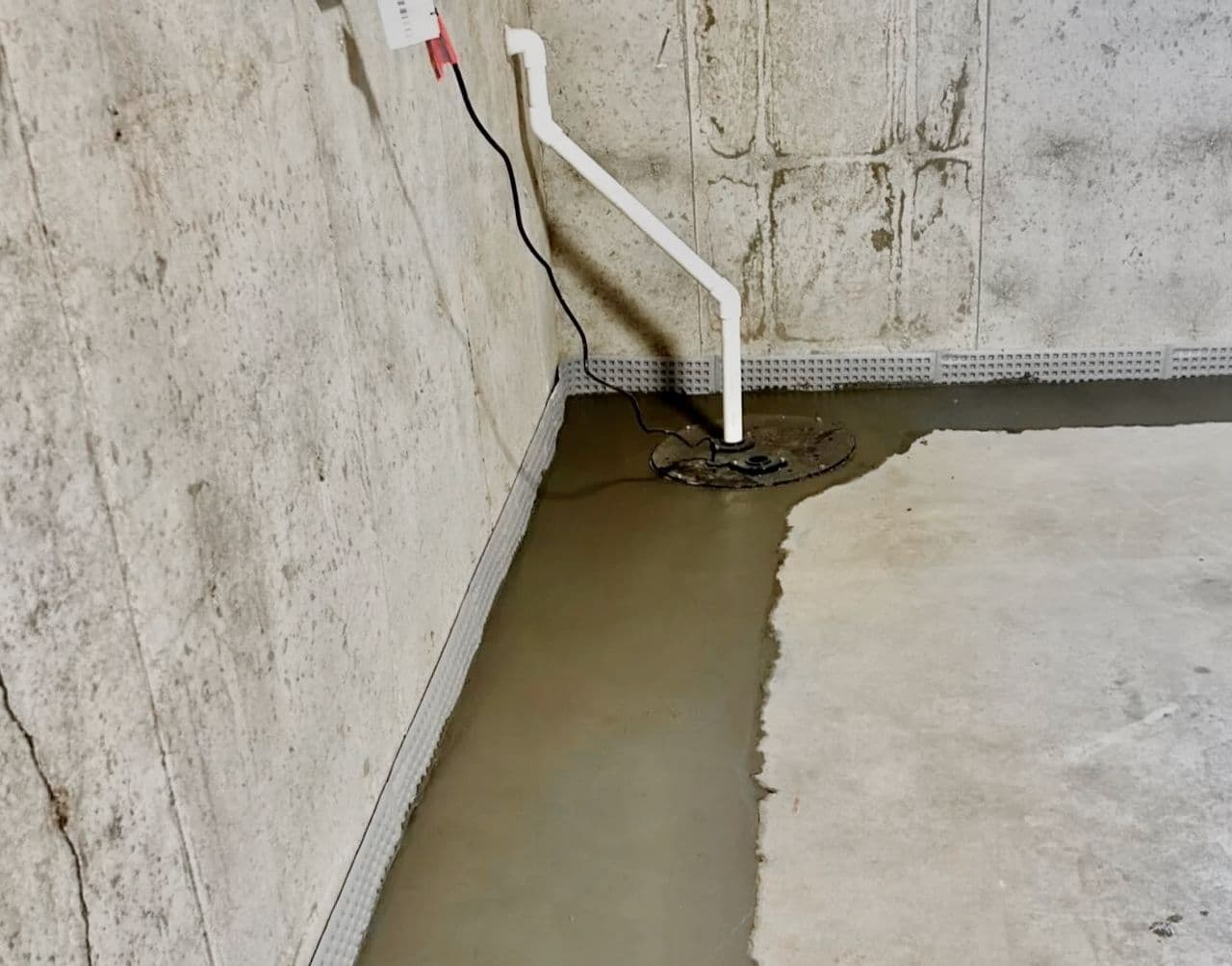 Basement sump pump basin with discharge pipe and drainage mat installed along a concrete block foundation wall
